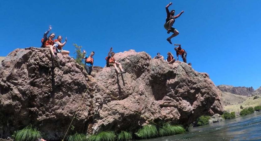 A student wearing safety gear jumps off a rocky ledge into a body of water. Others wearing safety gear sit on the ledge and watch.